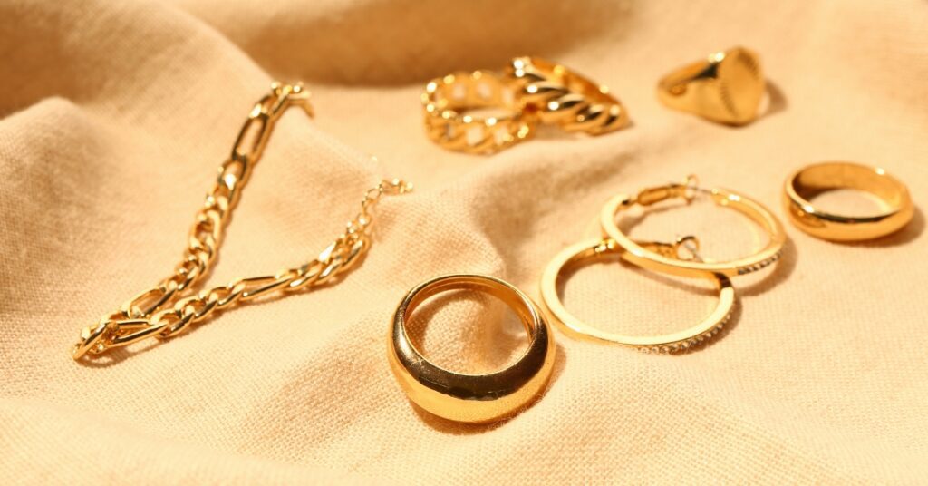 A collection of gold jewelry, including bracelets, rings, and hoop earrings, arranged on a beige fabric, gleaming in warm, natural light.