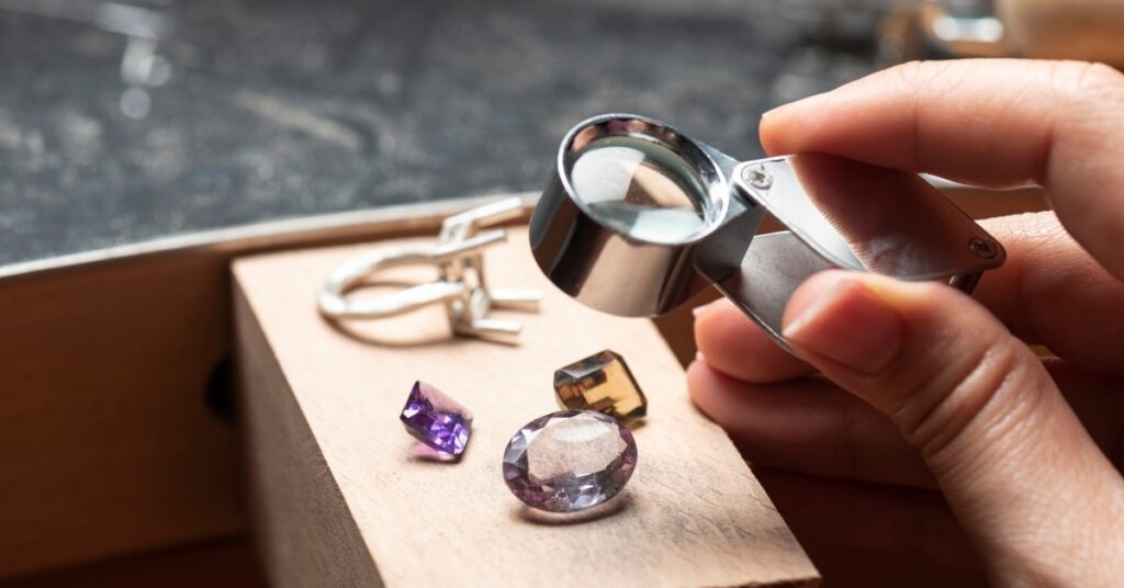 A jeweler examining various gemstones with a magnifying glass on a workbench, showcasing a ring setting and a collection of colorful gems.