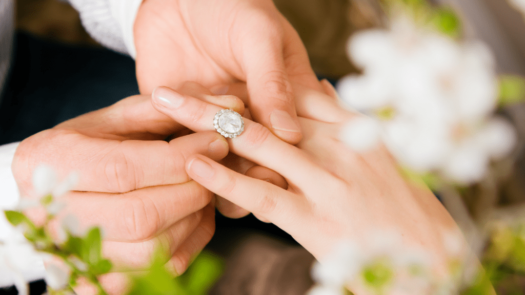 A man gently places a beautiful oval diamond engagement ring on his partner's finger amidst blooming flowers, symbolizing love and commitment.
