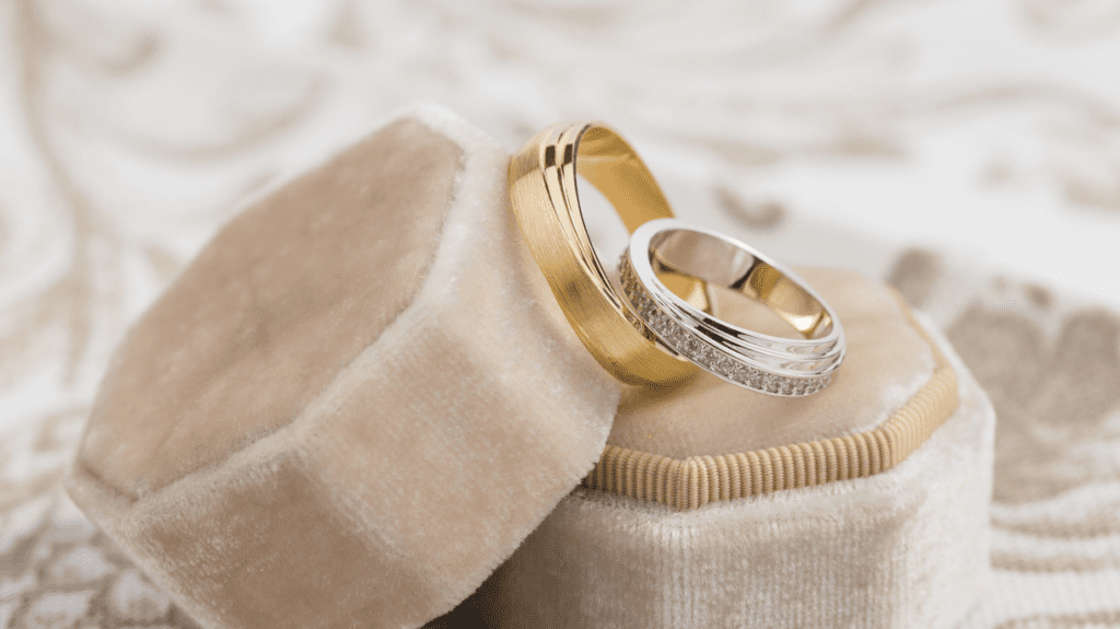 Two elegant wedding bands, one gold and one silver with diamonds, are displayed on plush velvet ring boxes against a luxurious, patterned background.