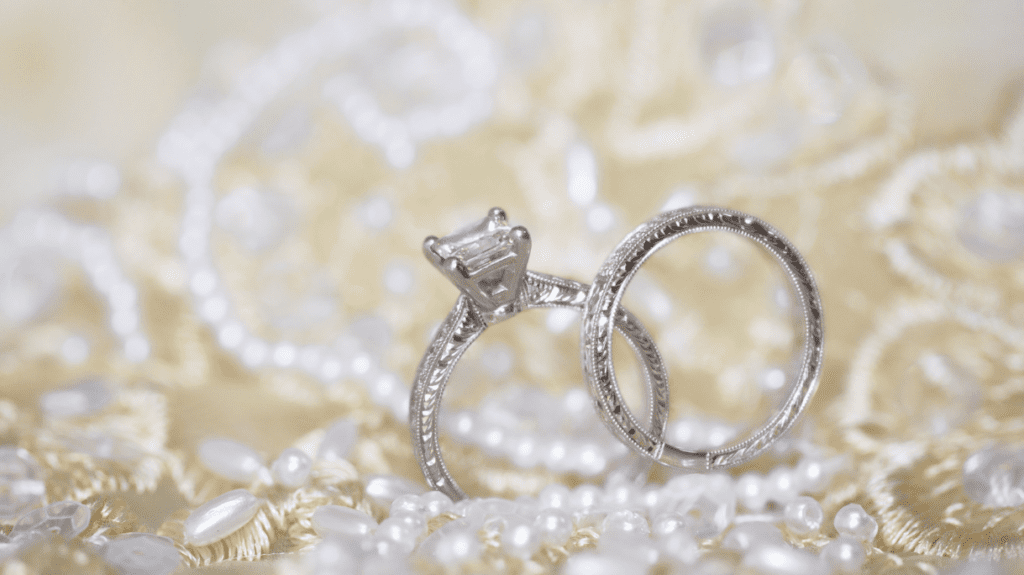 Intricately designed platinum engagement and wedding rings set against a backdrop of pearls and lace, showcasing timeless elegance and craftsmanship.