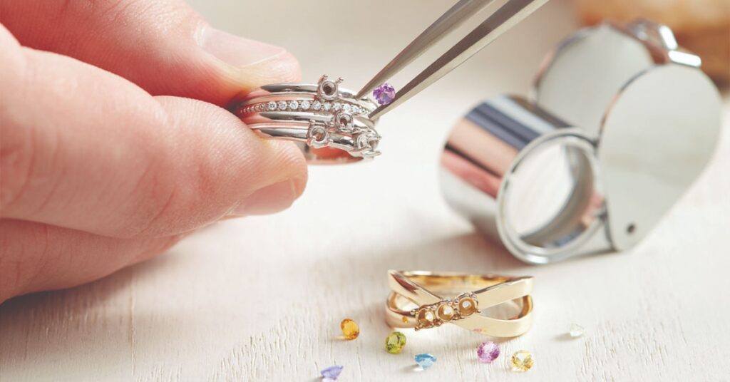 A jeweler's hand holding silver rings with tweezers, selecting a purple gem to place. Various gemstones and a magnifying loupe are in the background.