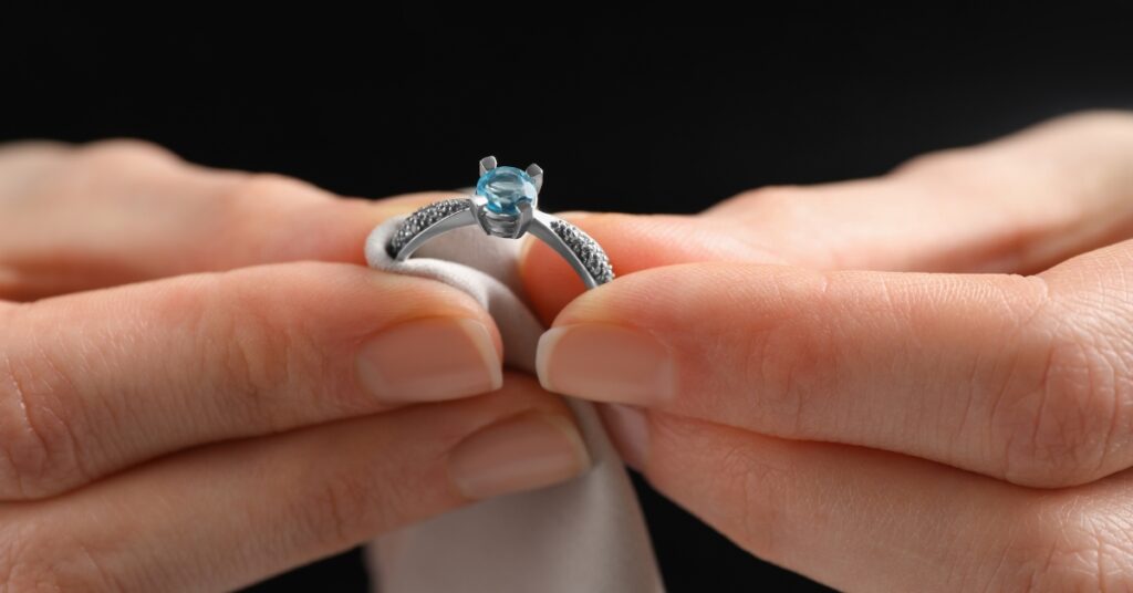 A person delicately holds a silver ring adorned with a blue gemstone, highlighting the intricate details and elegance against a dark background.