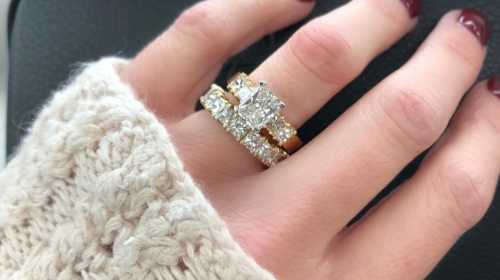 A hand wearing a cozy sweater displays a dazzling set of rings; one features a square-cut diamond, complemented by two eternity bands with round-cut diamonds.