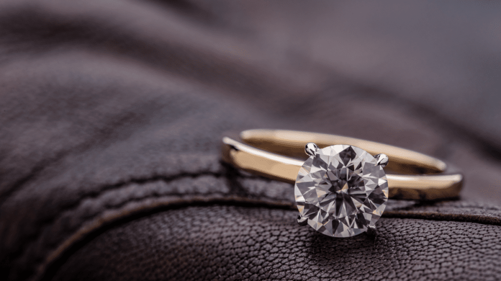 A close-up of a solitaire diamond engagement ring with a gold band, placed on a textured brown leather surface, showcasing its brilliant cut.