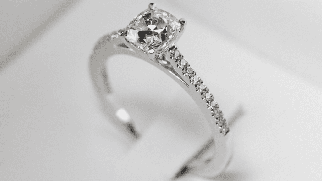 Elegant diamond engagement ring with a cushion-cut center stone and pavé diamond band, displayed against a sleek white background.