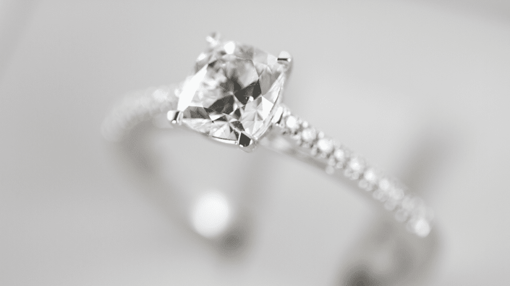 Close-up of a diamond engagement ring with a cushion-cut center stone and a pavé diamond band, elegantly displayed against a soft white backdrop.