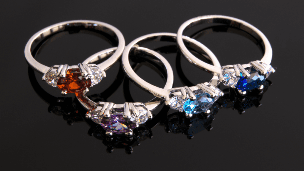 Four elegant silver rings with oval gemstones in red, purple, light blue, and dark blue, set with smaller white stones, reflecting on a glossy surface.