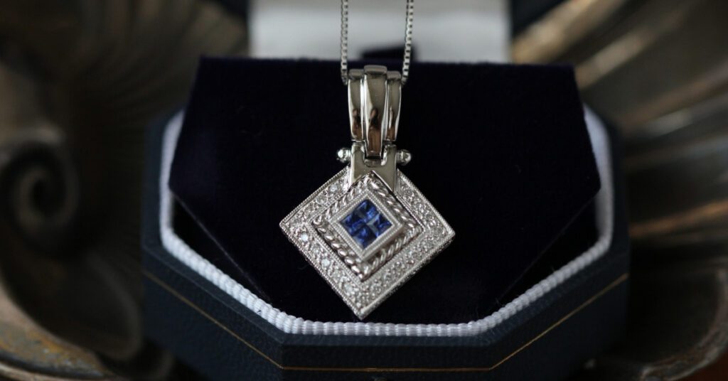 Elegant silver pendant with a square blue gemstone centerpiece, surrounded by intricate detailing, displayed in a dark velvet jewelry box.