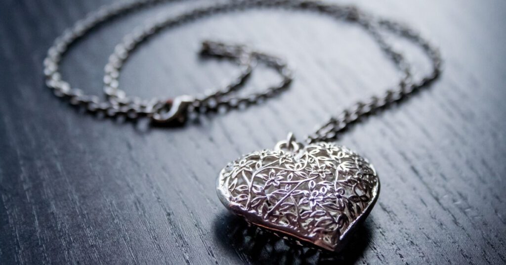 Intricately designed heart-shaped silver locket with floral patterns on a delicate chain, resting on a dark wooden surface, evoking vintage charm.