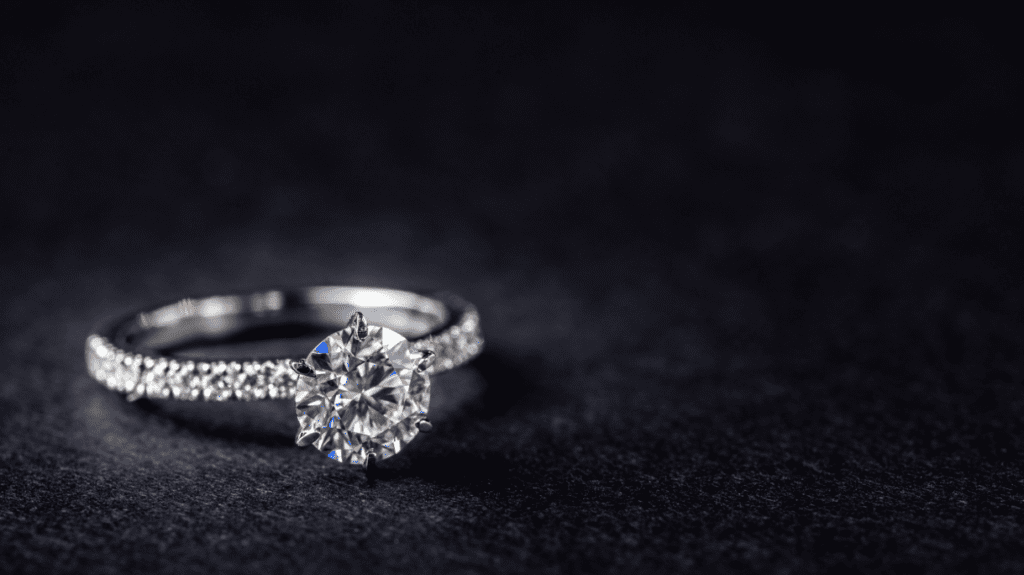 A solitaire diamond engagement ring with a round brilliant-cut center stone, set on a diamond-studded band, elegantly displayed on a dark surface.