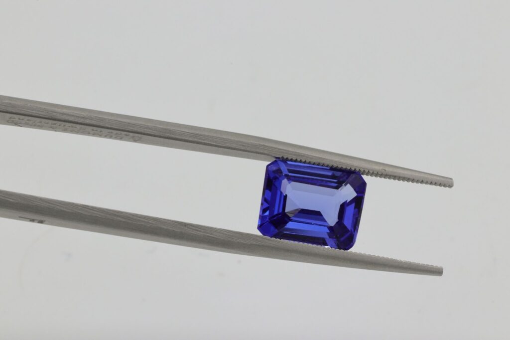 A pair of tweezers holding a radiant, rectangular-cut blue gemstone against a neutral background, emphasizing its deep color and precise facets.