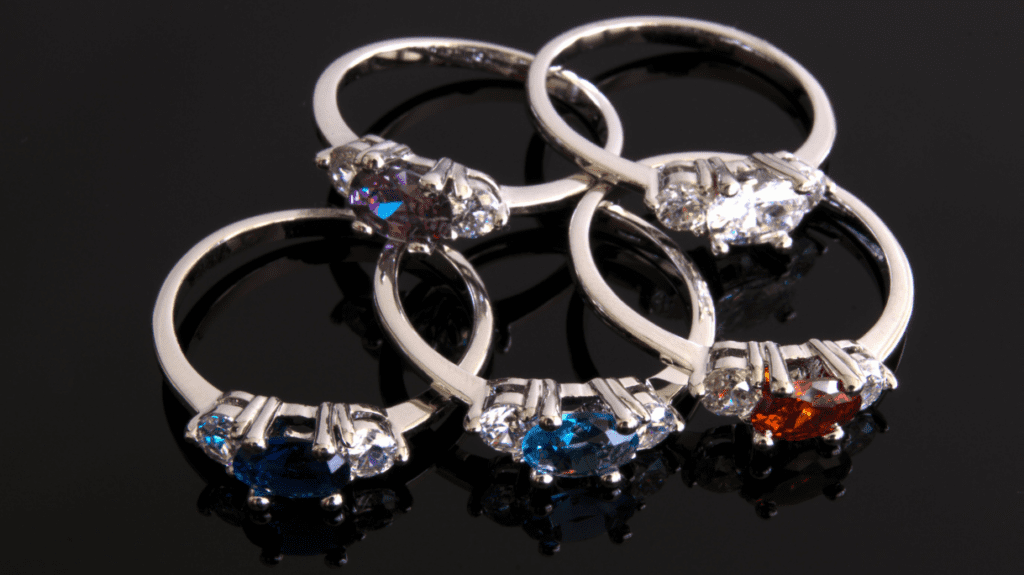 Four elegant silver rings with vibrant gemstones in purple, clear, blue, and red, arranged in a circle on a glossy black surface.