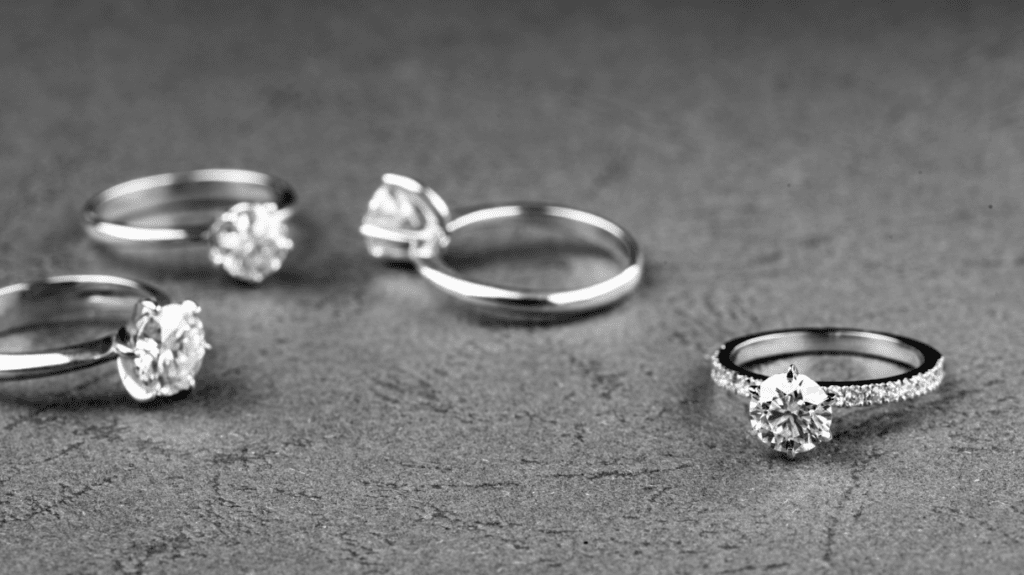 Four elegant diamond rings displayed on a textured surface, showcasing various settings and band styles in a sophisticated black and white shot.