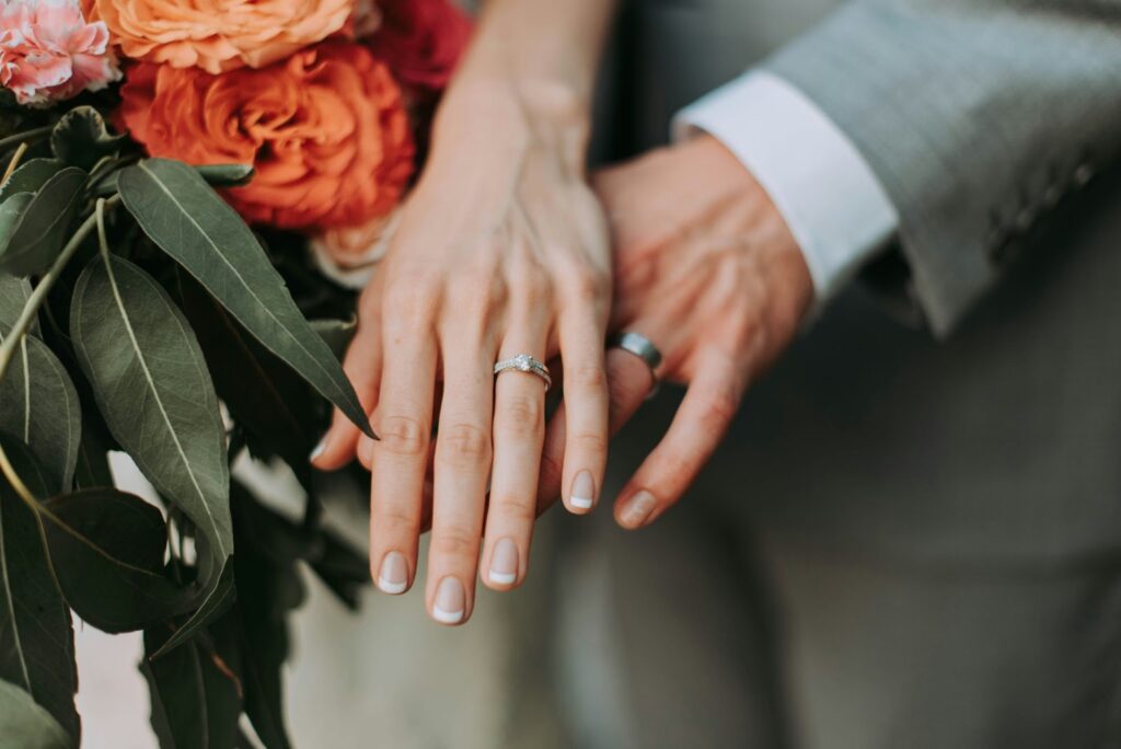 A close-up of a bride and groom's hands, showcasing their wedding bands against a backdrop of a vibrant floral bouquet.