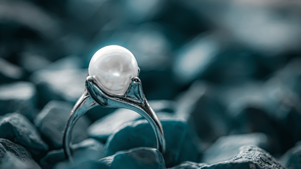 A close-up of a pearl ring with a silver band, elegantly displayed against a background of teal-colored stones, highlighting its intricate design.