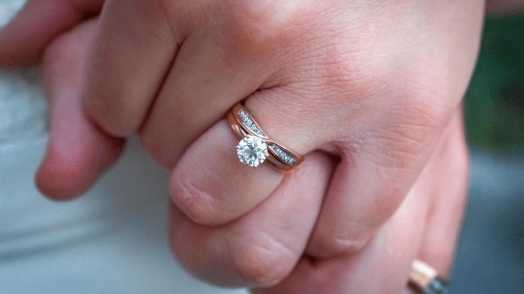 Close-up of a hand showing a gold engagement ring with a single round diamond and side stones, alongside a matching gold wedding band with diamonds.