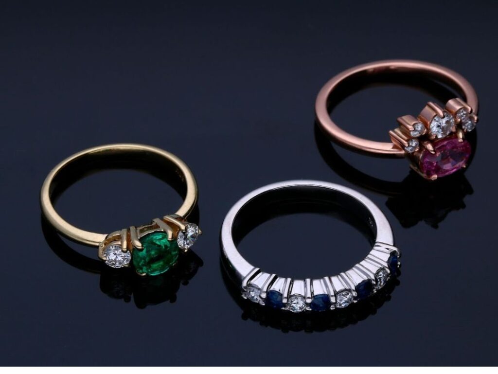 Three stunning engagement rings with emerald, pink sapphire, and sapphire gemstones set in yellow, rose, and white gold bands on a black background.