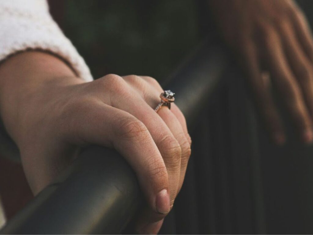 A close-up of a hand wearing an engagement ring, gripping a black railing, with another hand blurred in the background, suggesting a couple.