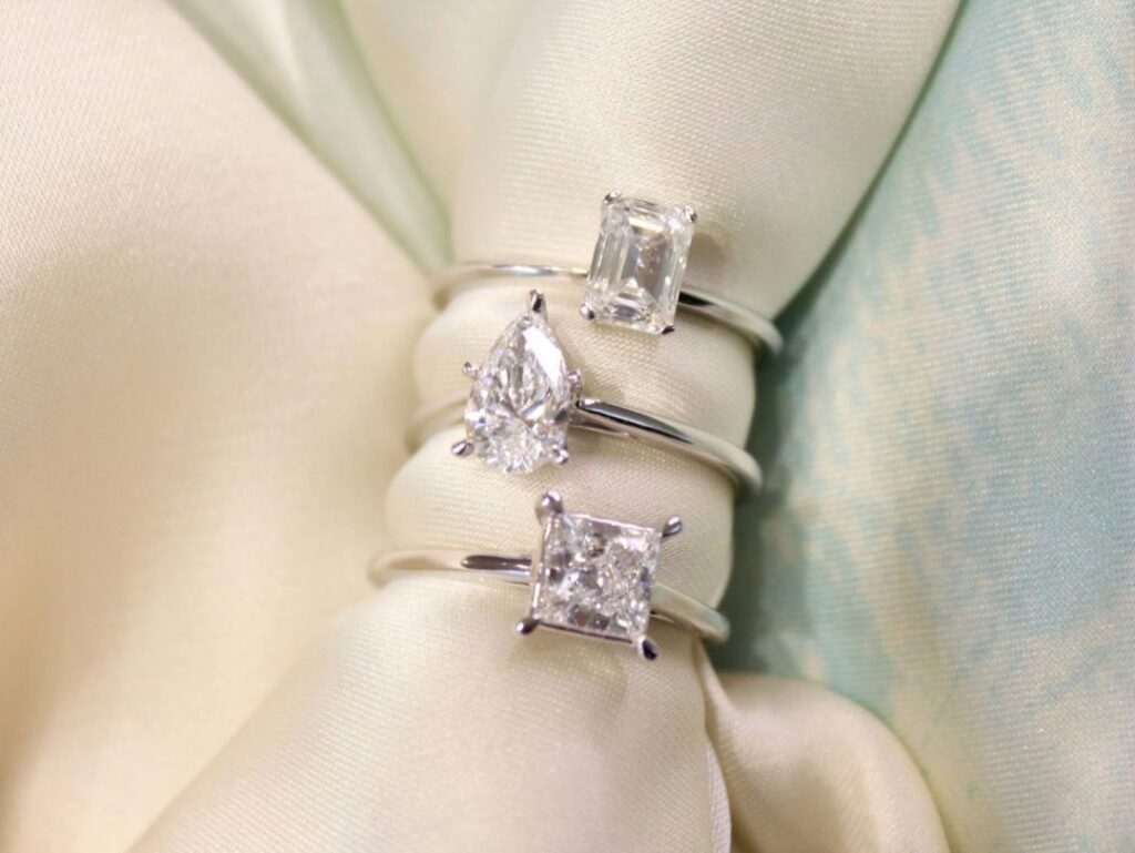Three diamond rings with different cuts (emerald, pear, princess) displayed on a soft, pale fabric background, highlighting their brilliance.