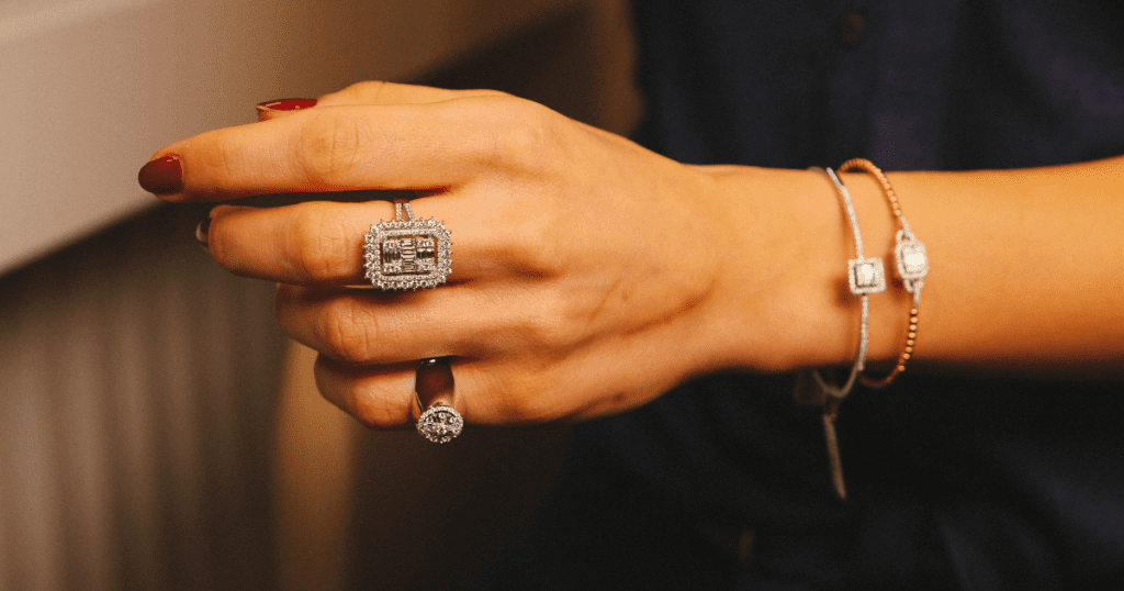 A hand with red nail polish adorned with a dazzling square diamond ring and a smaller round diamond ring, accompanied by elegant silver bracelets.