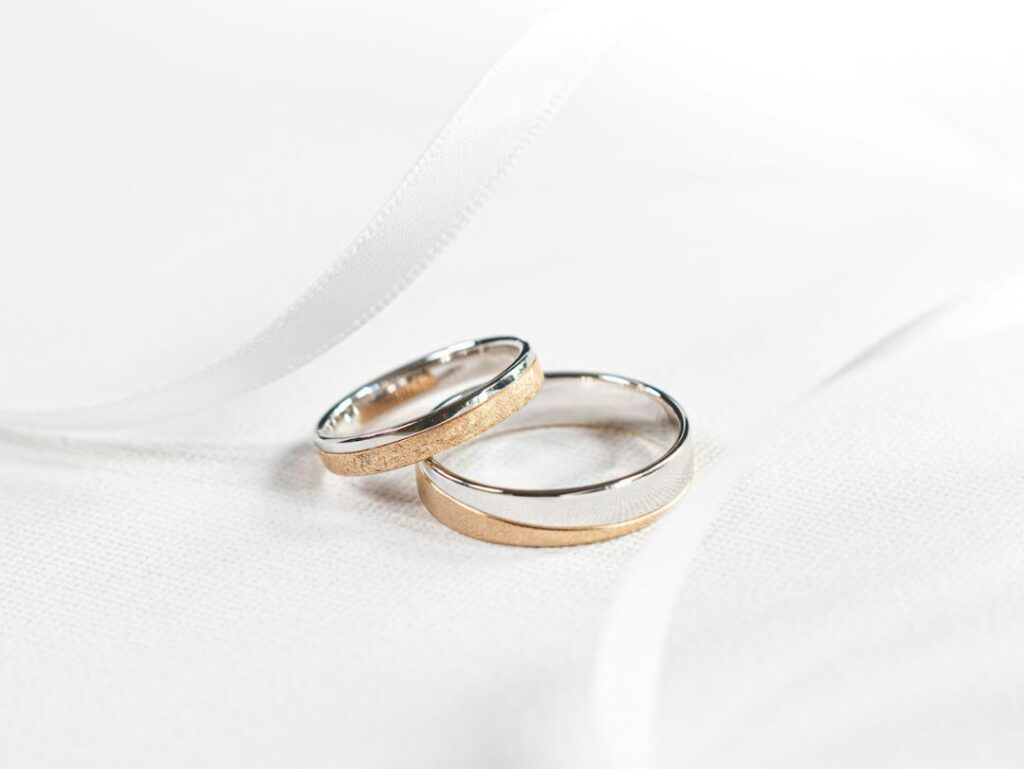 Two interlocking wedding bands with a mix of gold and silver, resting on a white fabric background with a ribbon softly curving around them.