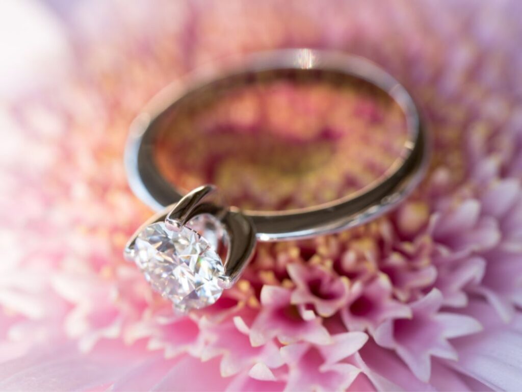 A close-up of a diamond engagement ring placed on a pink flower, highlighting the sparkle of the diamond against the soft floral petals.
