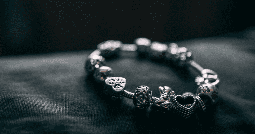 A silver charm bracelet with intricate heart and floral designs, delicately resting on a dark fabric surface, highlighted by soft, moody lighting.