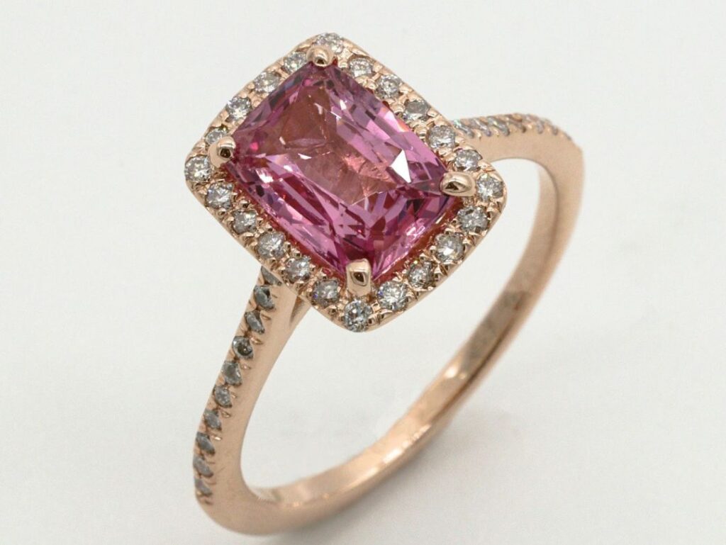A radiant-cut pink gemstone set in a rose gold ring, framed by a halo of small round diamonds, and a diamond-encrusted band for added sparkle.