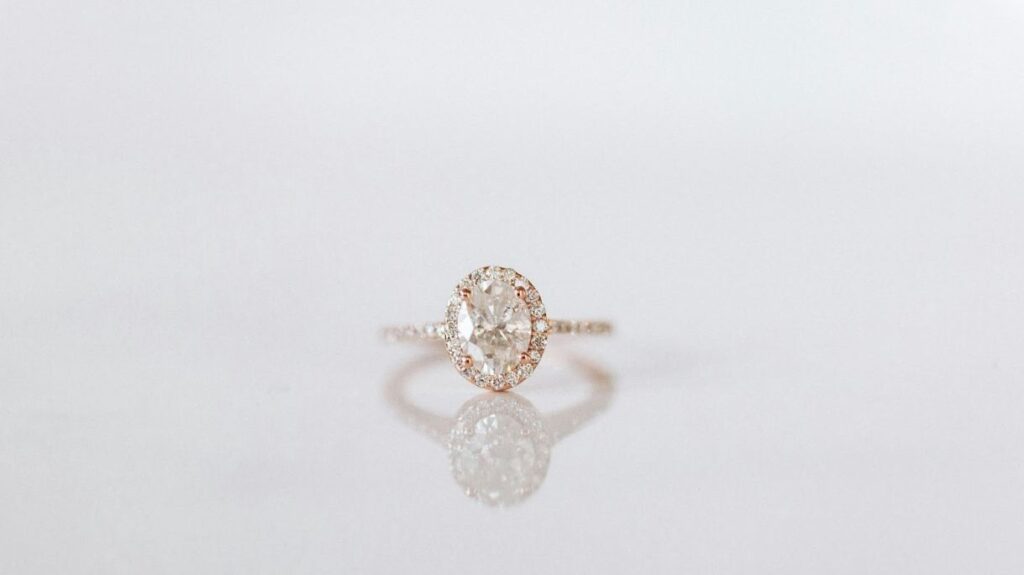 A delicate rose gold ring with an oval-cut diamond center, surrounded by a halo of smaller diamonds, featuring a thin, diamond-studded band.