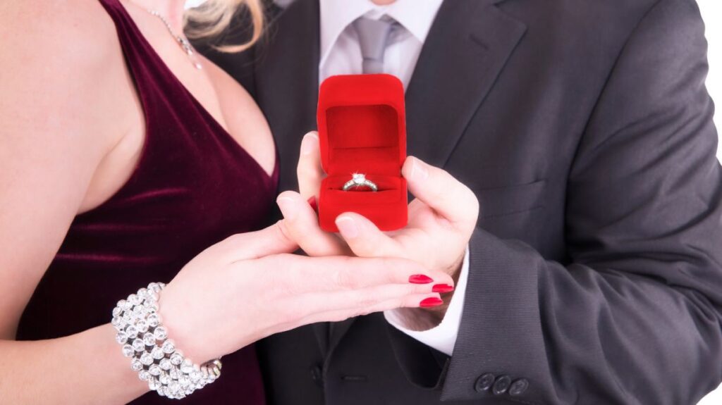 A couple dressed elegantly, with the man presenting a red velvet box holding a sparkling diamond ring, as the woman admires the gesture.