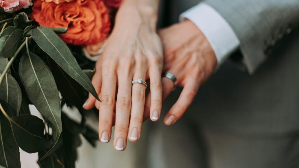 Newlywed couple's hands clasped, showcasing sparkling diamond engagement ring on bride's finger.