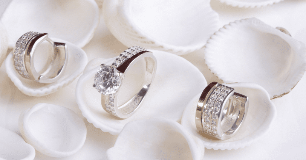 A close-up of a sparkling diamond ring and earrings set against a bed of seashells.