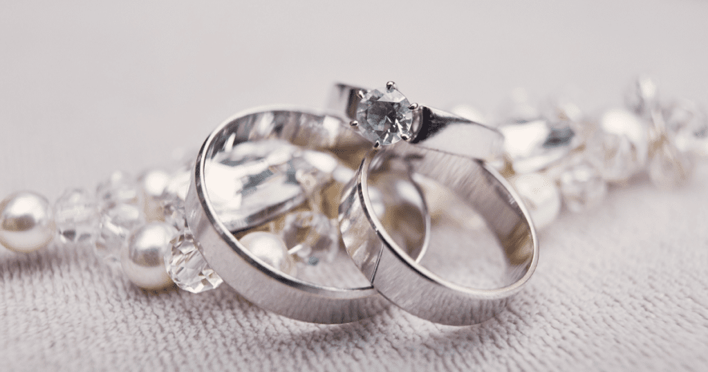 A close-up of intertwined wedding bands and an engagement ring with a sparkling diamond, resting on a bed of pearls and crystals.