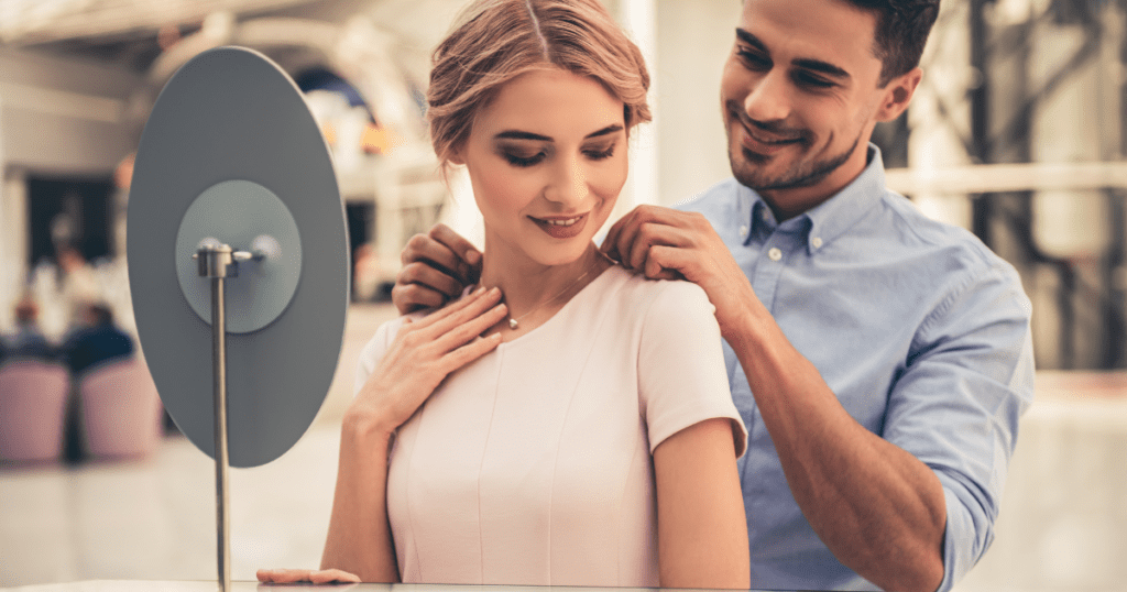 A smiling couple admires a necklace in a jewelry store; the man helps the woman put it on as she looks in the mirror, showcasing their happy moment.