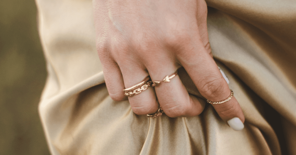 A close-up of a woman's hand adorned with delicate gold rings, gently clutching a soft beige fabric, showcasing minimalist elegance.