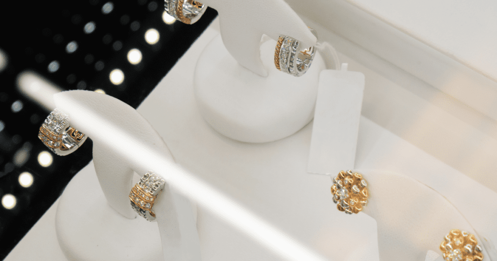 A close-up view of luxurious rings displayed on white stands, featuring intricate gold and diamond designs under bright showroom lights.