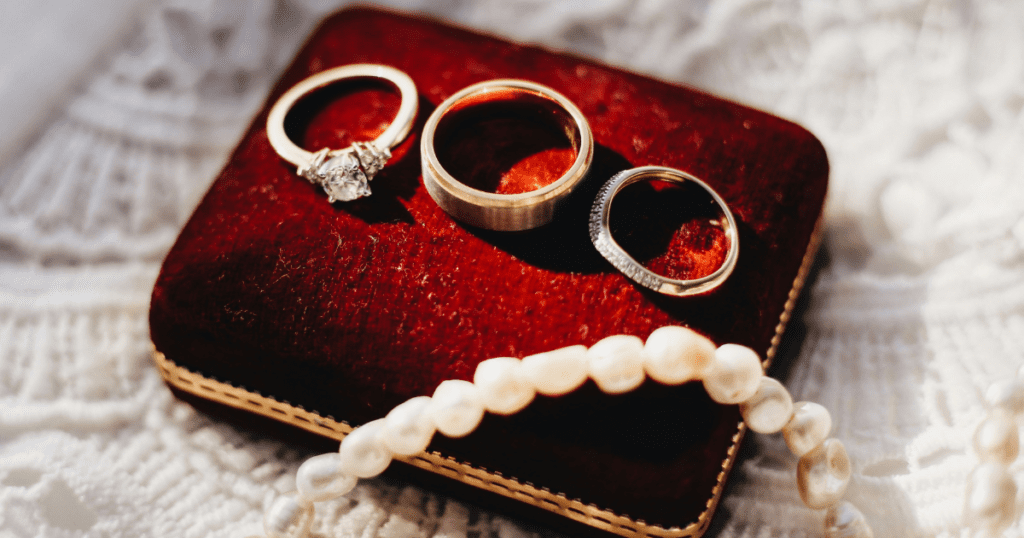 Three gold rings, including a diamond engagement ring, displayed on a plush red velvet box, accented with a strand of pearls.