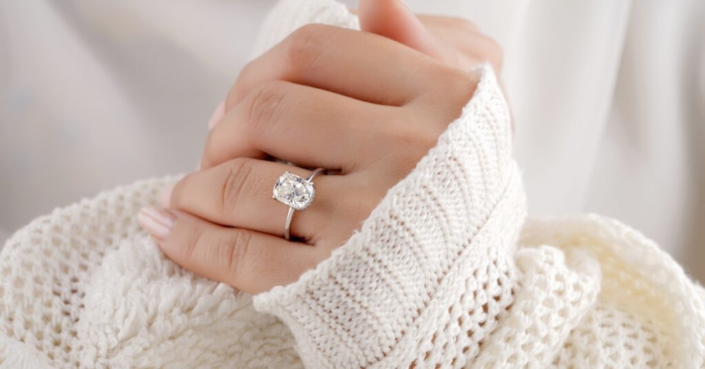 A hand gently holds a cozy white sweater, showcasing a sparkling diamond ring on the finger, highlighting elegance and warmth.