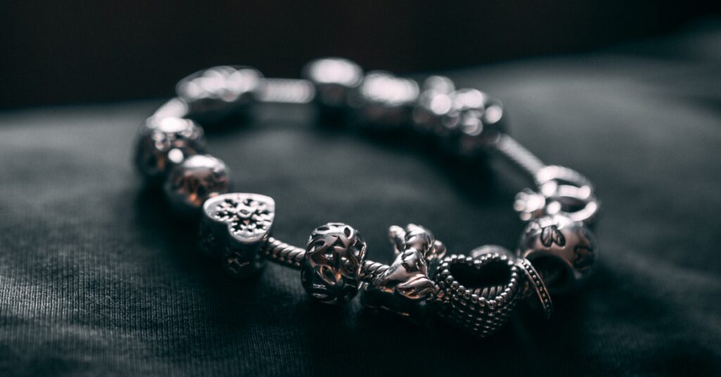 A silver charm bracelet rests on a dark surface, adorned with intricate charms including hearts, flowers, and a coiled snake design.