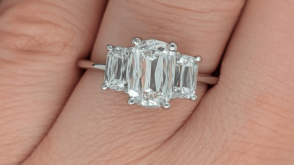  Elegant three-stone engagement ring with emerald-cut center diamond and smaller side stones, resting on a finger.