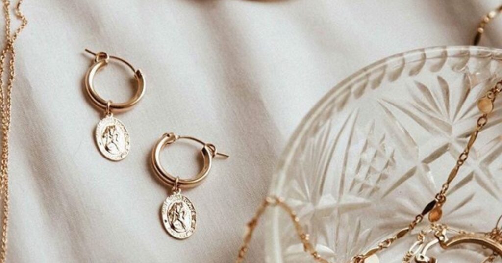 Gold hoop earrings featuring engraved coin charms, displayed on a soft white fabric next to delicate gold chains and a glass dish.