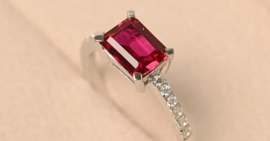 A radiant deep red emerald-cut gemstone set in a sleek silver band adorned with sparkling diamonds, showcased on a soft beige background.