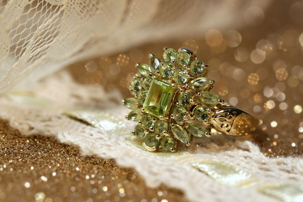 Elegant gold ring featuring a vibrant green gemstone surrounded by intricate smaller stones, placed on a delicate lace fabric with a glittery background.