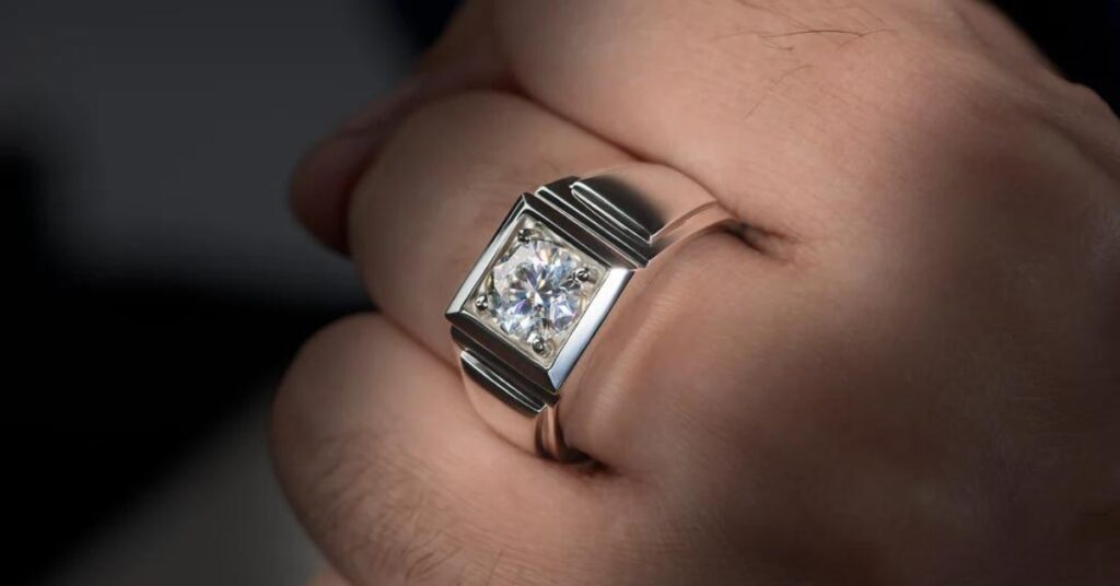 
A close-up of a hand wearing a sleek silver ring featuring a large, round-cut diamond securely set in a bold, geometric design.