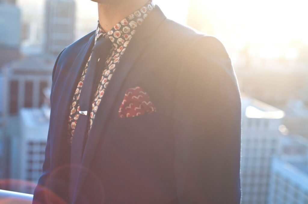 A man in a dark blue suit with a patterned shirt and tie, wearing a pocket square and tie clip.