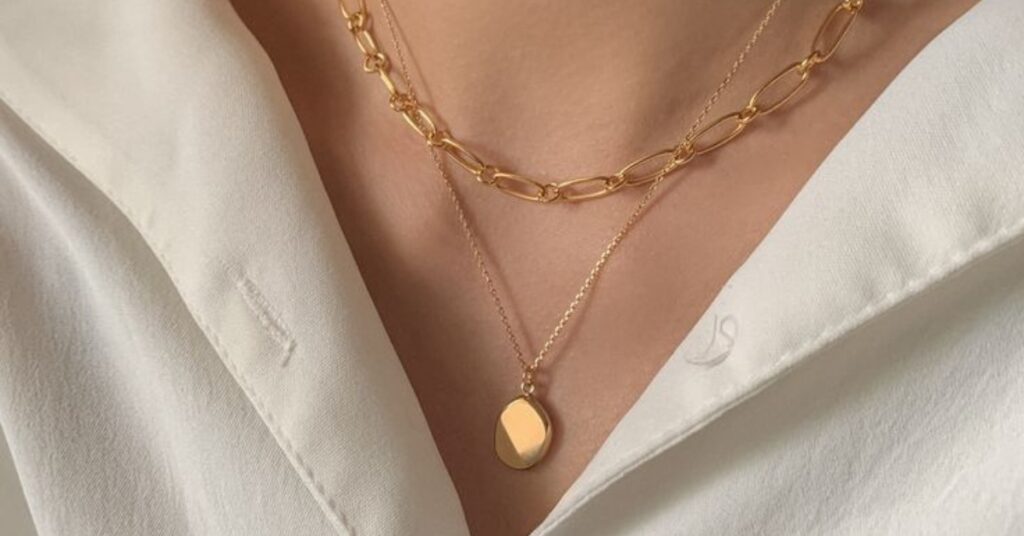 Close-up of a gold chain necklace with a linked design, paired with a second dainty necklace featuring a smooth oval pendant, worn over a white blouse.