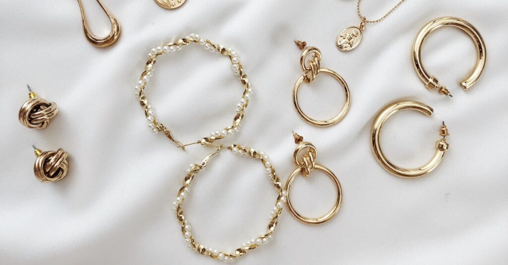 Assorted gold-tone jewelry on a white fabric background, including hoop earrings, pearl-accented hoops, knot studs, and a coin pendant necklace.