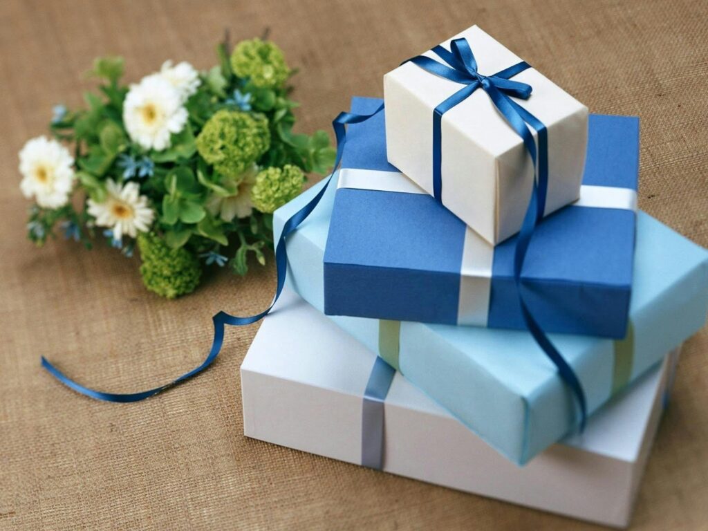 Stack of beautifully wrapped gifts in blue and white, tied with ribbons, next to a bouquet of flowers.