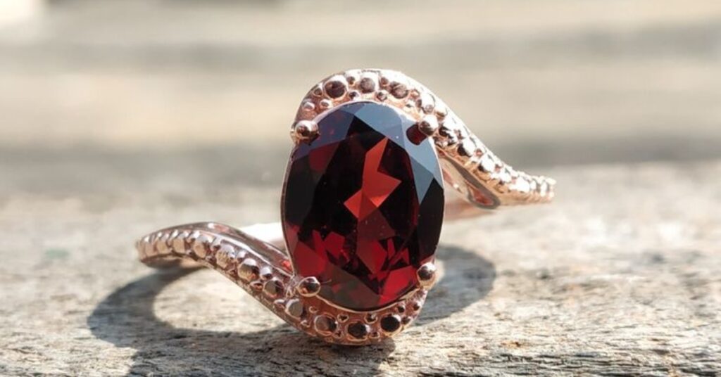 A striking deep red oval-cut gemstone set in a rose gold ring, with an intricate wave-like design and dotted accents along the band for added elegance.